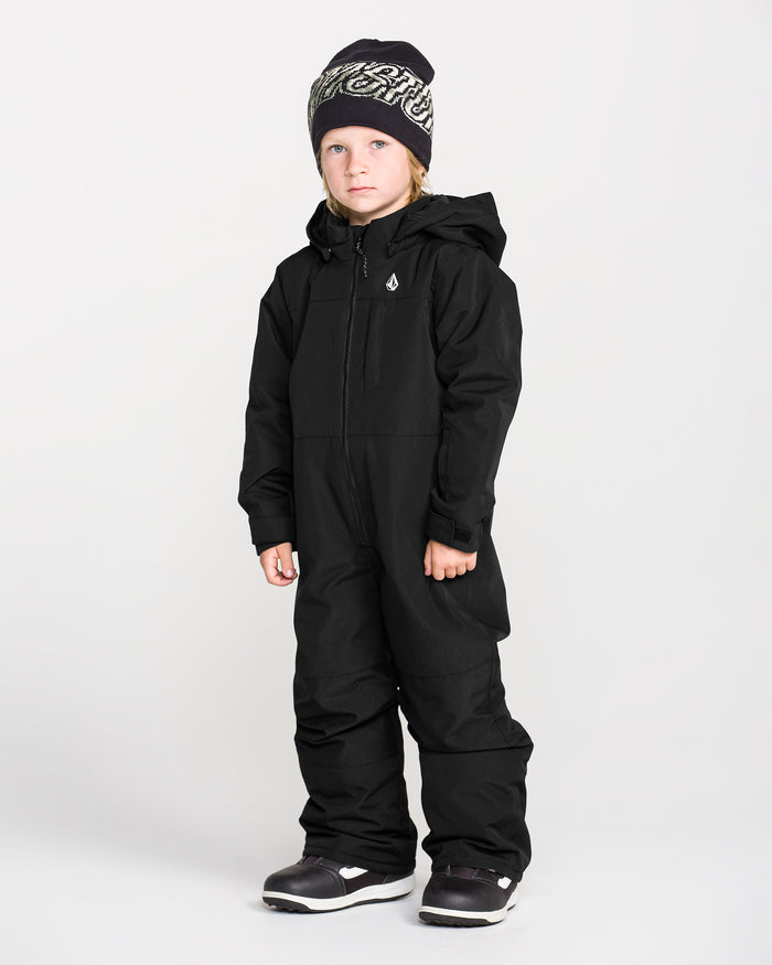 A young child stands against a plain white background wearing the Volcom Kids’ Little Little Flurry One Piece Snow Suit, paired with black winter boots and a black beanie with white lettering.