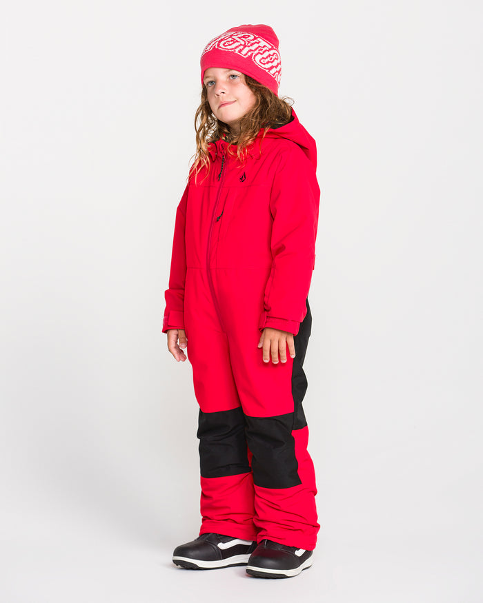 A child stands against a plain white background wearing the Volcom Kids’ Little Flurry One Piece in red—an insulated snowsuit featuring a hood, black knee patches, 10K/10K waterproof-breathable protection, and recycled V-Science fabric. A red beanie and black winter boots complete the cozy winter look.