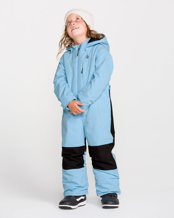 A child in the Volcom Kids’ Little Flurry One Piece, a light blue and black snowsuit crafted with recycled V-Science fabric, paired with black and white sneakers and a white beanie, stands against a plain background looking up and smiling.