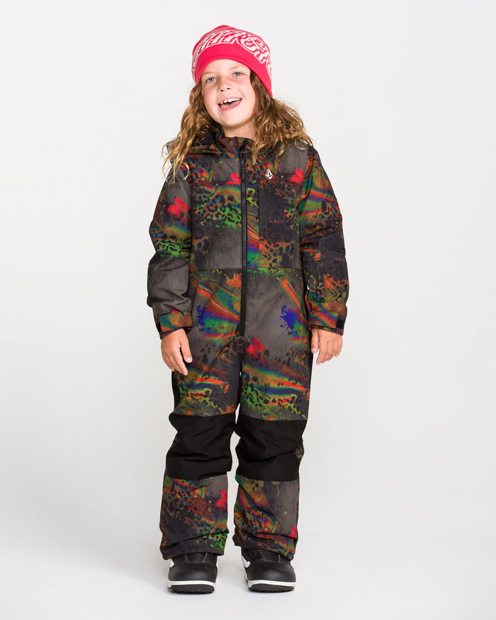 A smiling child wears the Volcom Kids’ Little Flurry One Piece, a colorful patterned snowsuit with warm insulation, paired with a pink beanie, while standing against a plain white background and looking slightly to the side.