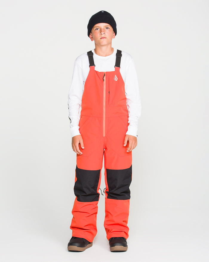 A young person wearing a black beanie, a white long-sleeve shirt, and the Volcom Kids’ Youth Barkley Insulated Bib in bright orange with black knee patches—designed with waterproof breathability and recycled insulation—stands facing forward against a plain white background.
