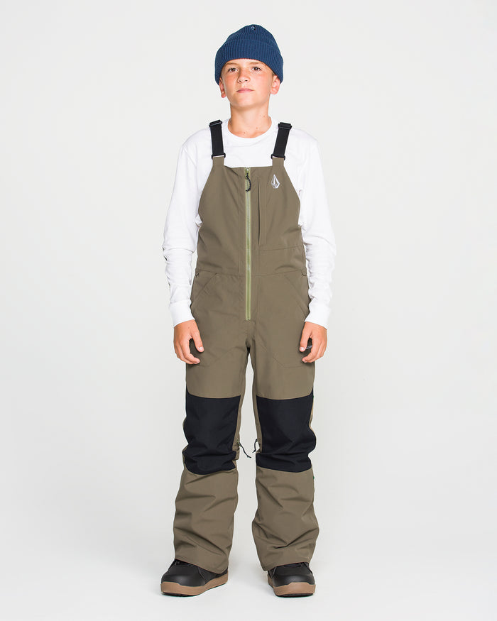 A boy wearing a blue beanie, white long-sleeve shirt, and Volcom Kids’ Youth Barkley Insulated Bib snow pants in olive green with black knee patches stands facing forward against a plain white background. The bib features waterproof insulation and Grow-Tech for lasting comfort and warmth.