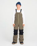 A boy wearing a blue beanie, white long-sleeve shirt, and Volcom Kids’ Youth Barkley Insulated Bib snow pants in olive green with black knee patches stands facing forward against a plain white background. The bib features waterproof insulation and Grow-Tech for lasting comfort and warmth.
