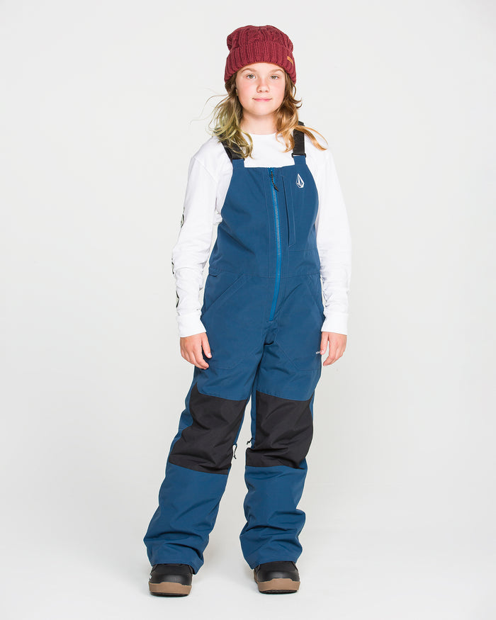 A child stands against a white background, wearing a red knit beanie, a white long-sleeve shirt, and Volcom Kids’ Youth Barkley Insulated Bib snow overalls in blue with black knee patches and brown boots. The overalls offer recycled insulation and waterproof breathability for all-day comfort and warmth.