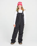 A young person wearing a pink beanie, white long-sleeve shirt, Volcom Kids’ Youth Barkley Insulated Bib in black with waterproof insulation, and brown winter boots stands facing forward against a plain white background.
