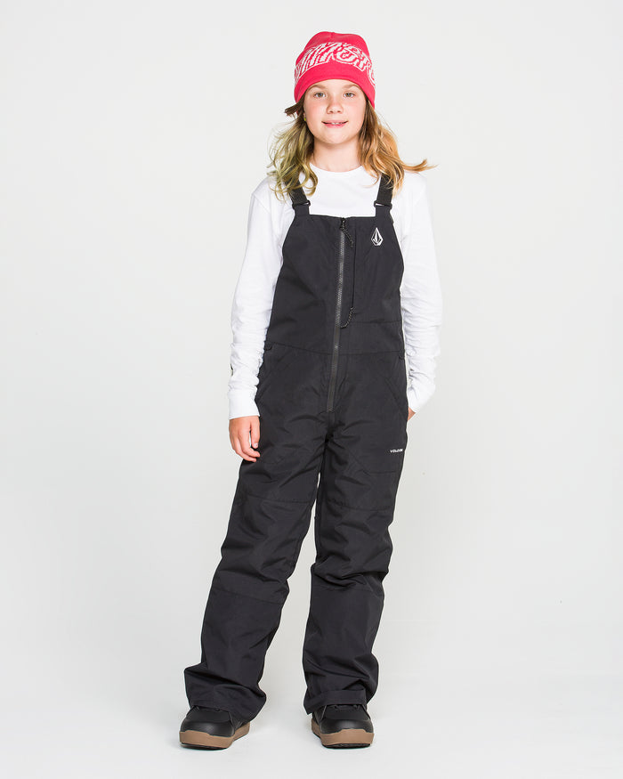 A young person wearing a pink beanie, white long-sleeve shirt, Volcom Kids’ Youth Barkley Insulated Bib in black with waterproof insulation, and brown winter boots stands facing forward against a plain white background.