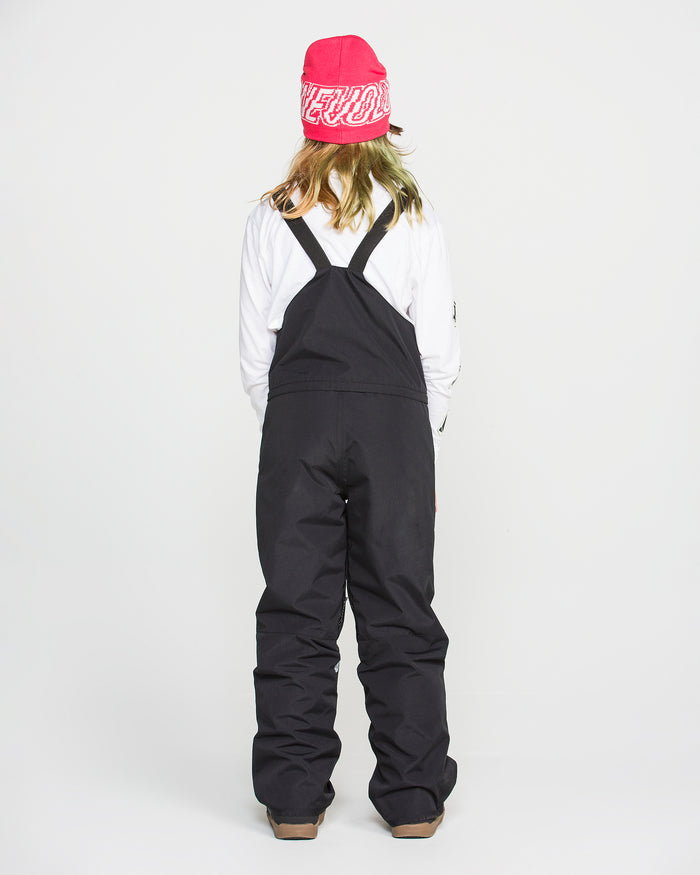A person stands facing away, wearing a pink beanie, white long-sleeve shirt, and the Volcom Kids’ Youth Barkley Insulated Bib—black snow overalls with straps featuring waterproof insulation—against a plain white background.