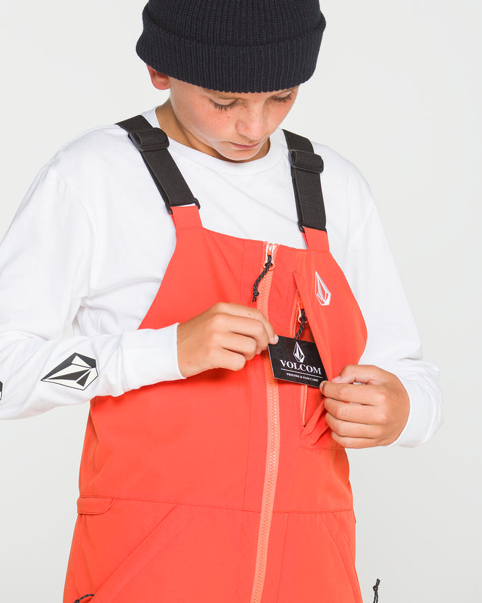 A person in a black beanie and white long-sleeve shirt opens the chest pocket of bright orange Volcom Kids’ Youth Barkley Insulated Bib overalls to reveal the Volcom tag, looking down at it and highlighting the style’s waterproof breathability.