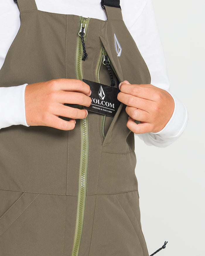 A person is shown wearing Volcom’s Kids’ Youth Barkley Insulated Bib in olive green over a white shirt, unzipping a pocket to reveal a black Volcom tag inside—highlighting the innovative Grow-Tech feature for adaptable sizing. Only the torso and hands are visible against a plain background.