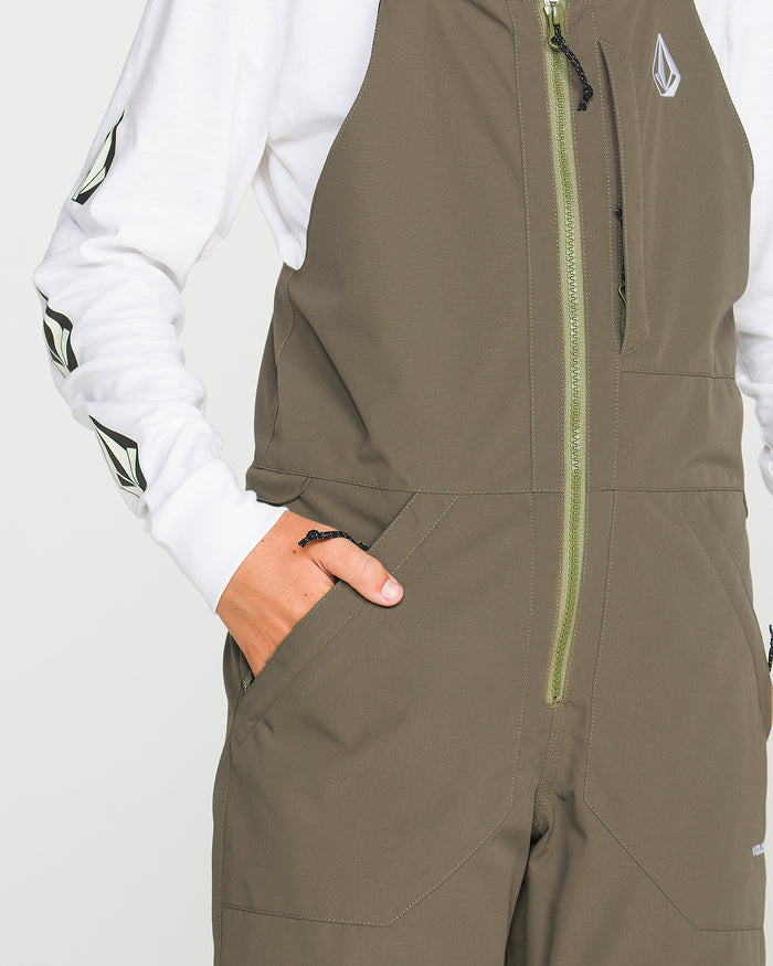 A person is wearing the Volcom Kids’ Youth Barkley Insulated Bib in olive green—an insulated, waterproof zip-up jumpsuit—layered over a white long sleeve shirt with sleeve logos. One hand is in their pocket, and the cropped image does not show their face.