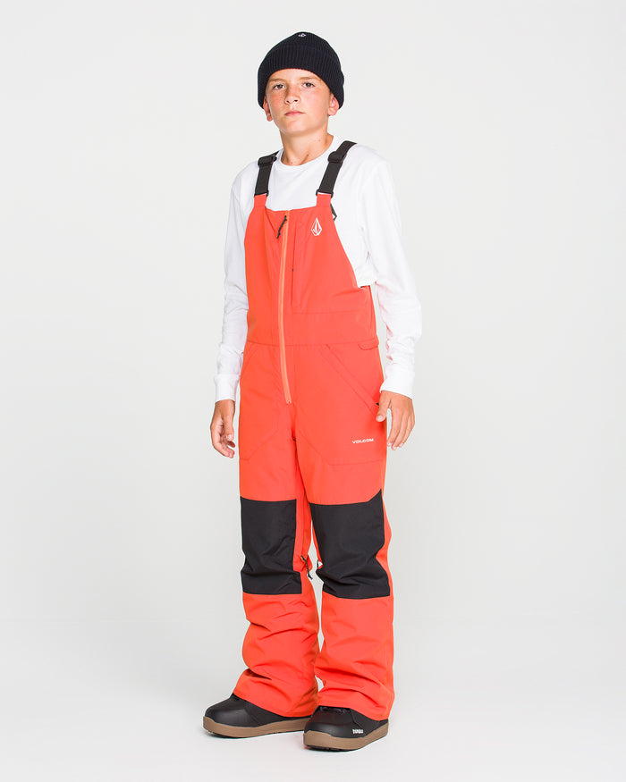 A young person stands facing forward against a plain white background, dressed in a black beanie, white long-sleeve shirt, and the Volcom Kids’ Youth Barkley Insulated Bib—bright orange snow pants featuring black knee patches and crafted for waterproof breathability.