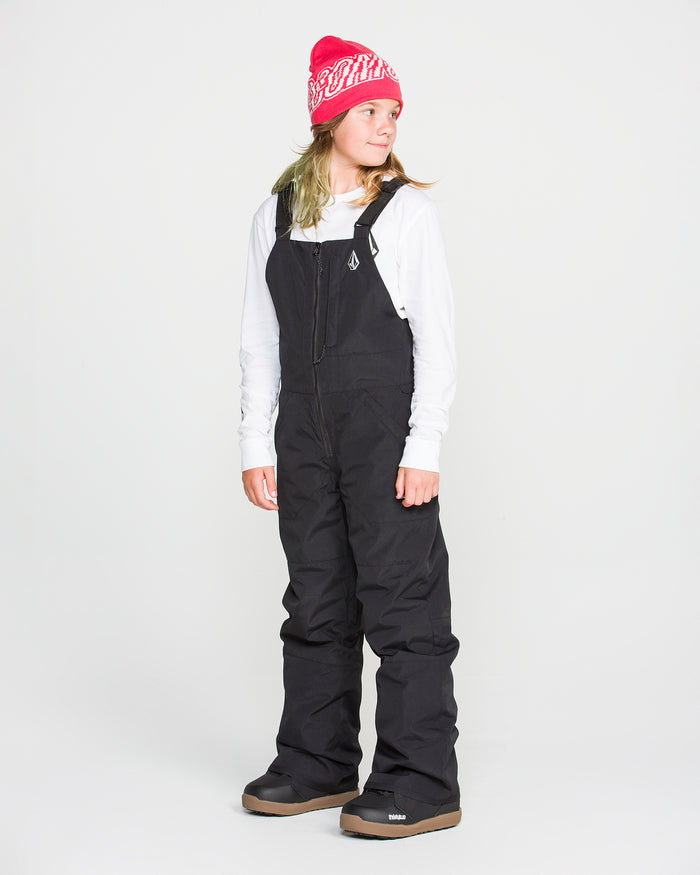 A person stands against a plain white background, looking to the side, dressed in a bright pink beanie, a white long-sleeve shirt, and the Volcom Kids’ Youth Barkley Insulated Bib—black snow overalls featuring waterproof insulation.