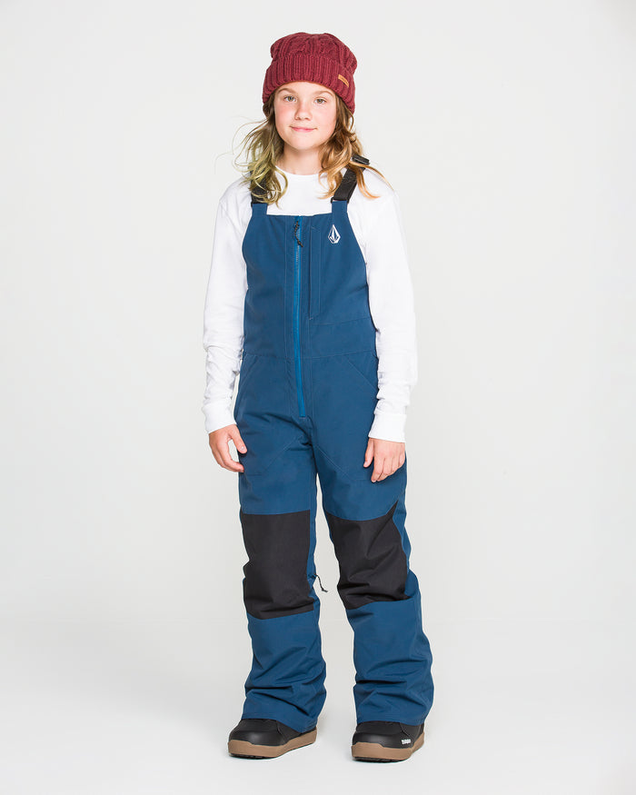 A child stands facing forward against a plain white background, wearing a red beanie, a white long-sleeve shirt, and the Volcom Kids’ Youth Barkley Insulated Bib in blue and black with recycled insulation.