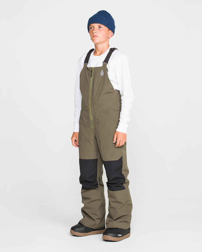 A young person in a blue beanie, white long-sleeve shirt, and olive green Volcom Kids’ Youth Barkley Insulated Bib stands against a plain white background.