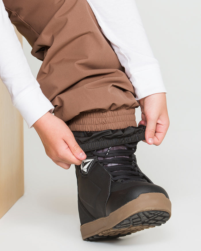 A person tucks their Volcom Kids’ Youth Chaos Insulated Pants, featuring elastic cuffs, into black and brown boots in preparation for outdoor activity. The V-Science 2-Layer construction and recycled insulation provide warmth and comfort throughout the adventure.