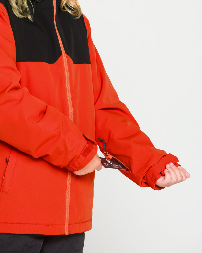 A person is shown from the torso down wearing the Volcom Kids’ Youth Stone.91 Insulated Jacket in red and black, holding a tag attached to the sleeve against a plain white background. Only their hands and torso are visible.