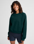 A smiling woman with dark wavy hair wearing a dark forest green crewneck sweatshirt and navy blue athletic shorts, standing against a light gray studio background.