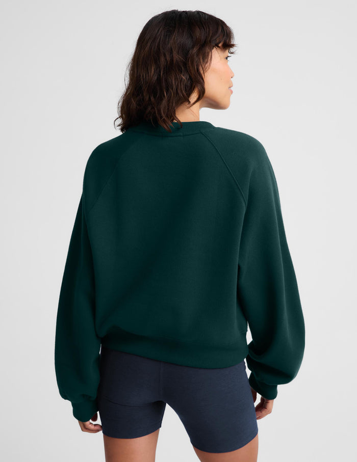 A woman shown from the back wearing a dark forest green crewneck sweatshirt and navy blue bike shorts against a light gray studio background.