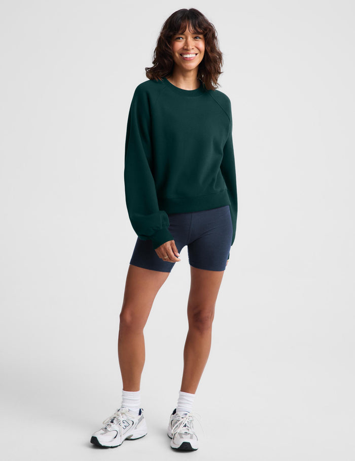 A woman modeling a dark green crewneck sweatshirt and navy blue athletic shorts against a light gray studio background.