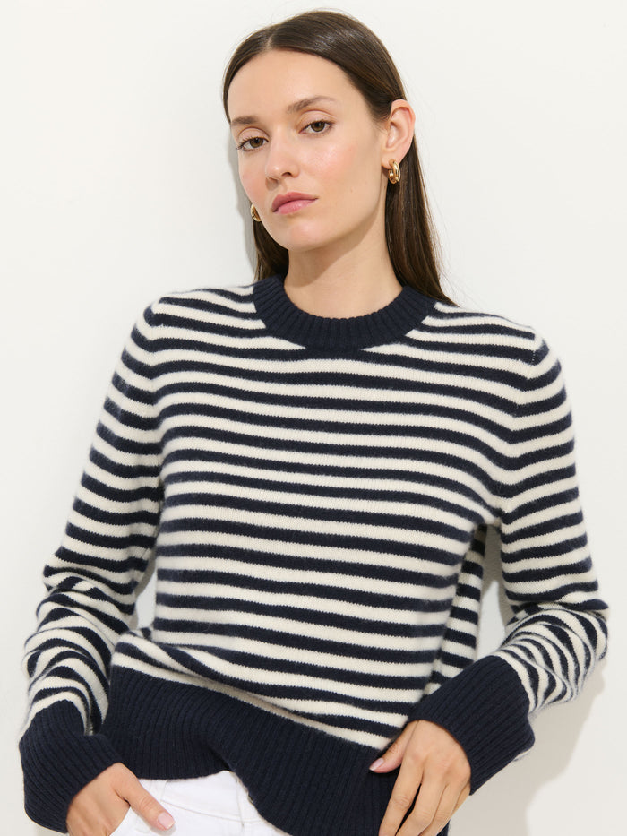 A woman, dressed in the Marais Striped Sweater in Washed Cashmere—a navy blue and white striped luxury knit—pairs it with white pants while standing against a plain white background. She has straight brown hair and gazes at the camera with a neutral expression.