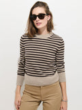 A woman stands against a plain white background, wearing the Marais Striped Sweater in Washed Cashmere—a luxurious black and beige striped piece crafted from 100% cashmere for exceptional softness. She pairs the sweater with tan pants and dark sunglasses, one hand resting in her pocket and a slight smile on her face.