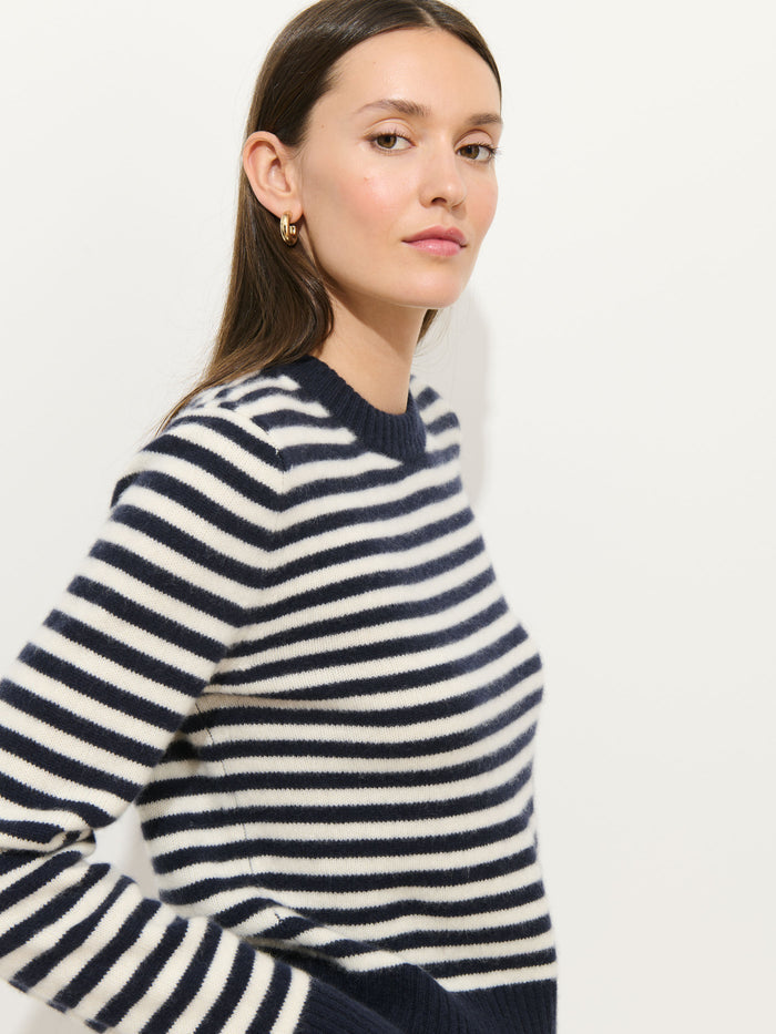 A woman with straight brown hair wears gold hoop earrings and the Marais Striped Sweater in Washed Cashmere, featuring navy and white stripes, while standing against a plain white background.