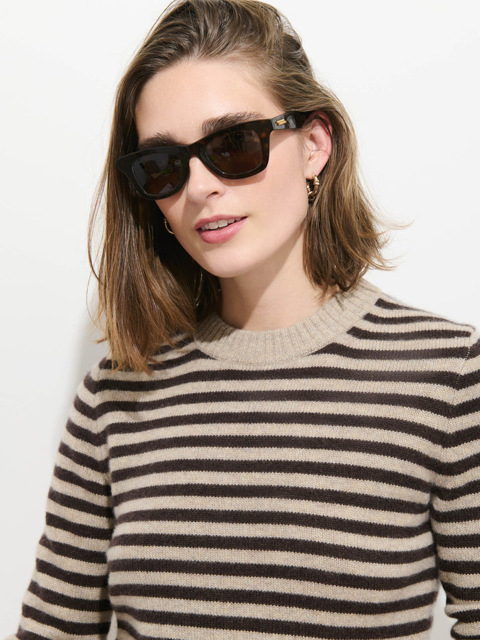 A woman with shoulder-length brown hair wears black sunglasses, gold hoop earrings, and the Marais Striped Sweater in Washed Cashmere—a beige and brown luxury sweater crafted from 100% cashmere for exceptional softness—while standing in front of a plain white background.