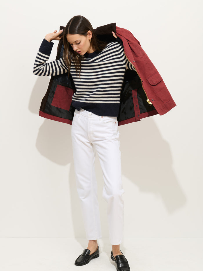A woman wearing the Marais Striped Sweater in Washed Cashmere, featuring a navy and white stripe pattern, pairs it with white pants and black loafers as she puts on a red jacket while looking down, standing against a plain white background.