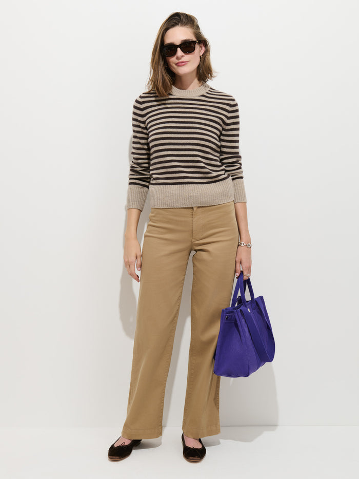 A woman in sunglasses wears the Marais Striped Sweater in Washed Cashmere—crafted from 100% cashmere for unmatched softness and an elevated look—with brown stripes and khaki pants, holding a large purple bag against a white background.
