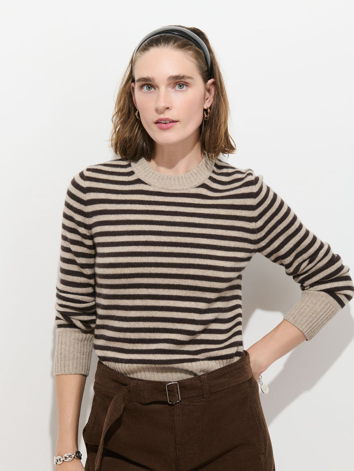 A woman with light brown hair wears a black headband, the Marais Striped Sweater in Washed Cashmere—a luxury sweater known for its extra softness—and brown pants. She stands against a white background with one hand on her hip, looking at the camera with a neutral expression.
