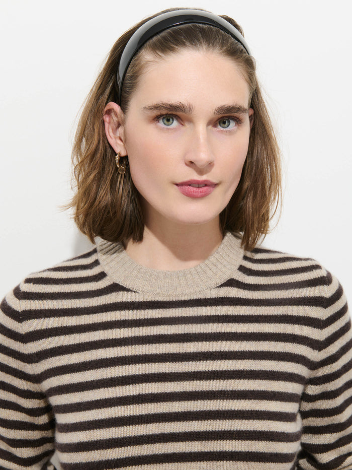 Wearing the Marais Striped Sweater in Washed Cashmere—renowned for its extra softness and featuring beige and black stripes—a person with shoulder-length brown hair, accented by a black headband and gold earrings, poses against a plain white background.