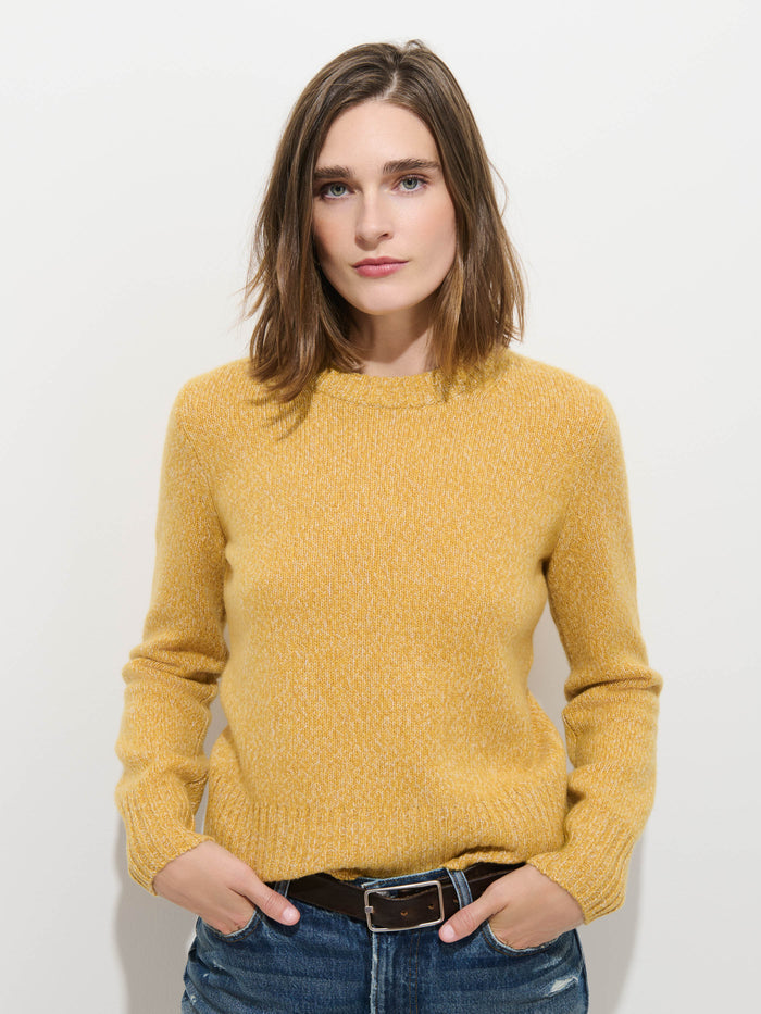 A woman with shoulder-length brown hair stands against a plain white background, her hands in her pockets, effortlessly highlighting the extra softness of the luxurious Marais Sweater In Cashmere—a premium 100% cashmere yellow knit—paired with blue jeans.