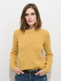 A woman with shoulder-length brown hair stands against a plain white background, her hands in her pockets, effortlessly highlighting the extra softness of the luxurious Marais Sweater In Cashmere—a premium 100% cashmere yellow knit—paired with blue jeans.