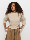 Wearing the Marais Sweater In Cashmere, a woman with curly hair and a brown skirt stands against a white wall, looking to the side with her hands on her hips, radiating luxury and extra softness.