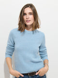 A woman with shoulder-length brown hair wears the Marais Sweater In Cashmere—a light blue, 100% cashmere piece known for its exceptional softness and luxury—paired with dark jeans. She stands against a white wall with her hands in her pockets, smiling softly at the camera.