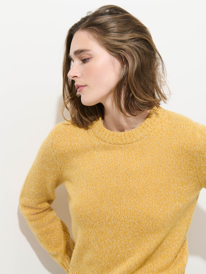 A person with shoulder-length brown hair wears the Marais Sweater In Cashmere, a yellow luxury sweater made from 100% cashmere, and looks to the side against a plain white background.
