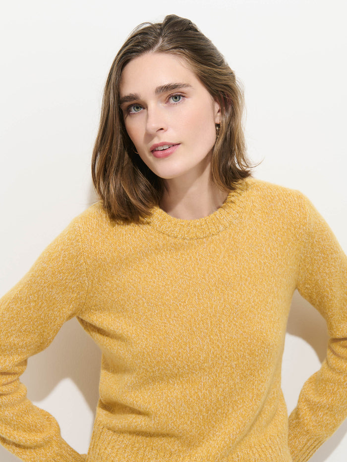 A person with shoulder-length brown hair stands against a plain white background, looking slightly to the side with a neutral expression while wearing the Marais Sweater in Cashmere—a luxurious yellow sweater crafted from 100% cashmere.