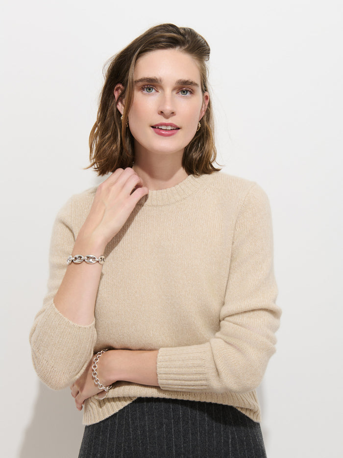 A woman with shoulder-length brown hair wears the Marais Sweater In Cashmere in beige, paired with gray pants. She smiles softly while standing against a plain white background, one hand gently touching her neck and the other arm crossed, radiating an air of exceptional softness and luxury.