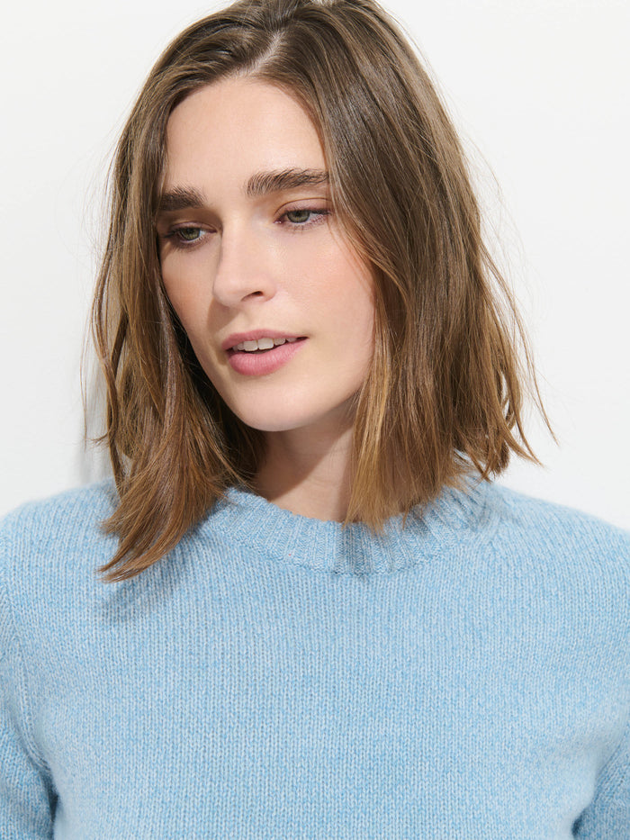 A woman with shoulder-length brown hair wears the Marais Sweater in Cashmere—a light blue, 100% cashmere piece renowned for its extra softness—and is posed against a plain white background, looking slightly downward with a neutral expression.