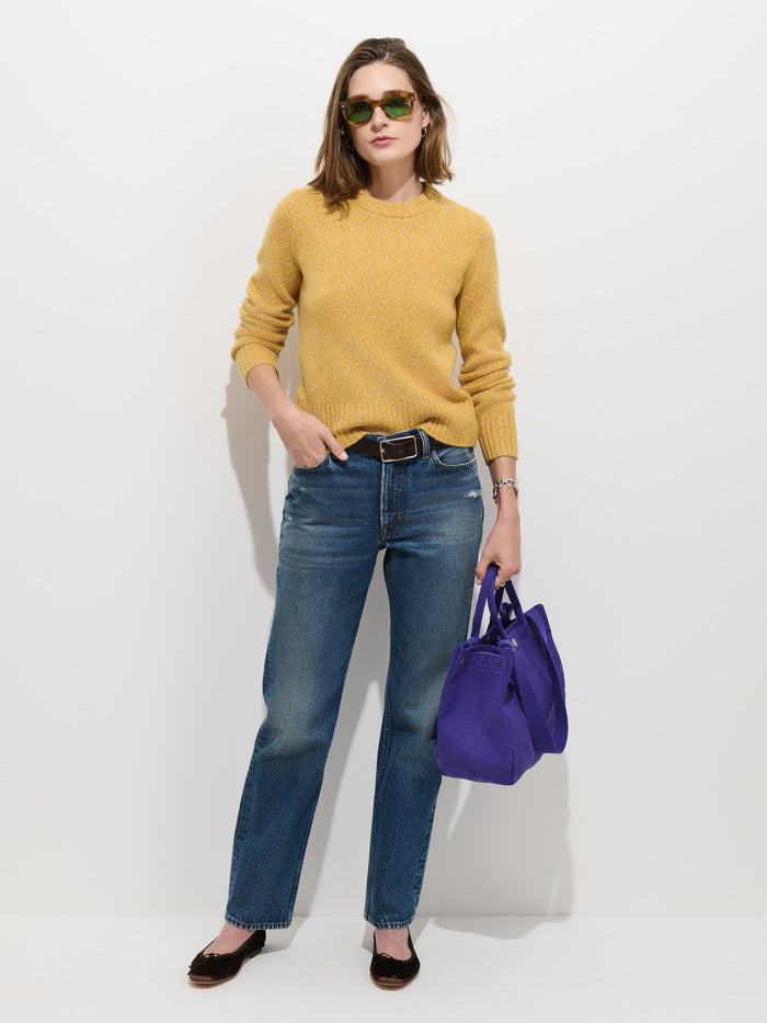 Woman in sunglasses wearing the Marais Sweater In Cashmere—a yellow luxury sweater crafted from 100% cashmere for extra softness—paired with blue jeans and black shoes, holding a bright purple handbag, standing against a plain white background.
