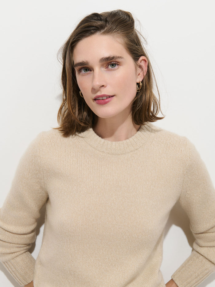 A woman with shoulder-length brown hair wearing the Marais Sweater in Cashmere stands against a plain white background, looking at the camera with a slight smile. This 100% cashmere piece exudes luxury and extra softness, offering a comfortable yet elegant look.