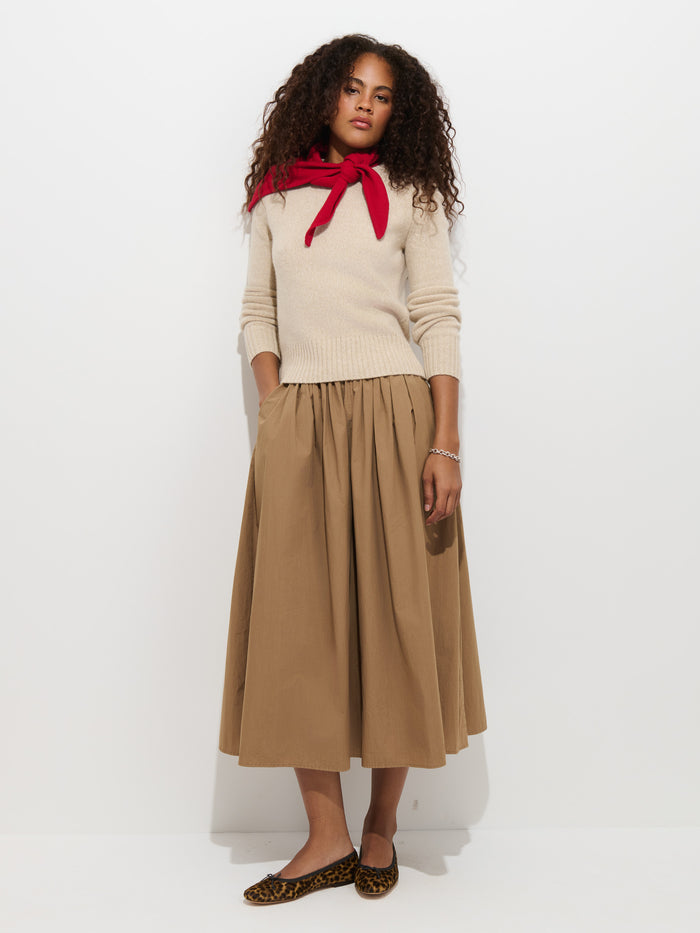 A woman wearing the Marais Sweater in Cashmere—renowned for its extra softness and luxury—in a cream shade, with a red sweater draped over her shoulders, paired with a tan pleated midi skirt and leopard print flats, stands against a white background.