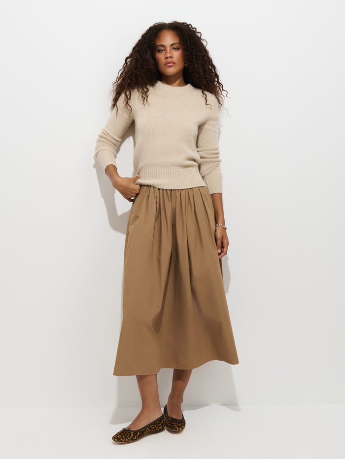 A woman with curly hair wears the Marais Sweater In Cashmere, paired with a tan midi skirt and leopard print flats, standing against a plain white background.