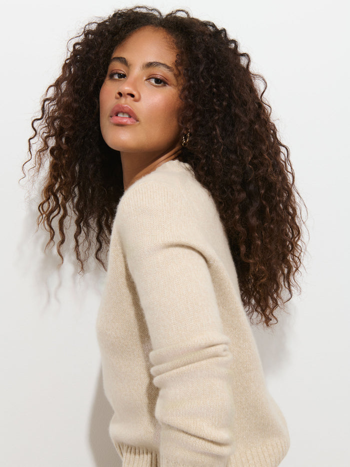A person with curly brown hair, dressed in the Marais Sweater In Cashmere in light beige, poses against a white background, looking confidently at the camera and exuding extra softness and luxury.