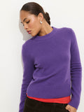 A woman wears the Marais Crewneck Sweater in Cashmere in purple over a red top with dark pants, standing against a light background, looking down with her hair pulled back and wearing small hoop earrings.