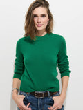 A person with shoulder-length brown hair wears the Marais Sweater in Washed Cashmere—a luxurious 100% cashmere green sweater—paired with blue jeans, standing against a plain white background with hands in pockets, looking at the camera.
