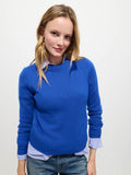 Wearing the Marais Sweater in Washed Cashmere—a bright blue luxury piece made from 100% cashmere for extra softness—a person with long blonde hair stands against a plain white background. The sweater is layered over a blue and white striped shirt and paired with blue jeans, as they smile slightly at the camera.