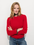 A blonde-haired woman stands against a white background, arms crossed and smiling slightly, wearing blue jeans and the Marais Sweater in Washed Cashmere—a bright red luxury sweater made from 100% cashmere—accented by a floral scarf around her neck to highlight its extra softness and elegance.