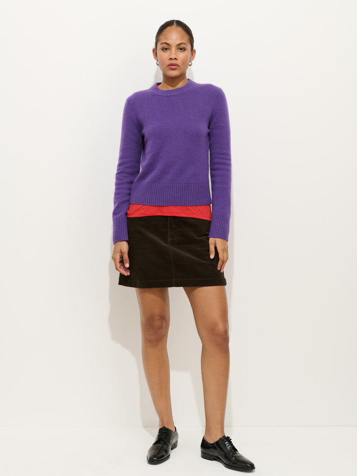 A woman stands against a white background wearing the Marais Crewneck Sweater In Cashmere in purple over a red top, paired with a dark brown skirt and black oxford shoes. She looks straight at the camera with a neutral expression.