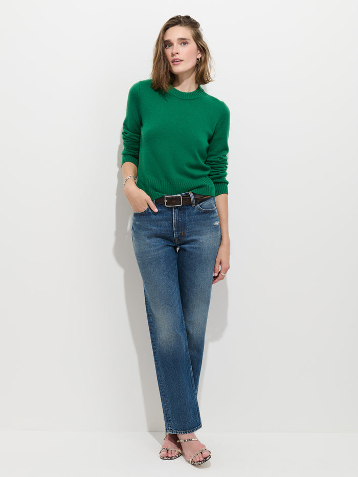 A woman wearing the Marais Sweater in Washed Cashmere—a luxurious, 100% cashmere green piece—paired with blue jeans and sandals stands against a plain white background, one hand in her pocket and the other resting by her side. The extra-soft texture of her sweater elevates her effortlessly stylish look.